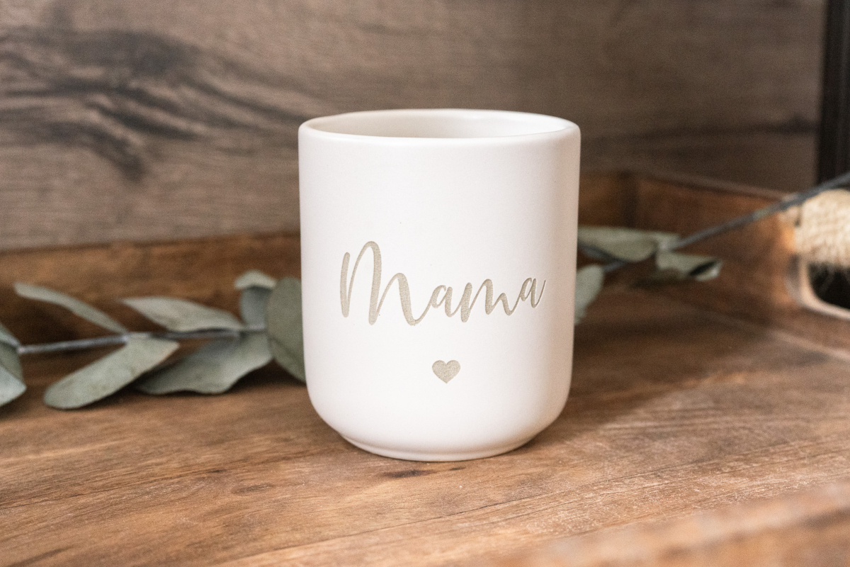 Matte weiße Keramiktasse mit Gravur „Mama“ und kleinem Herz auf Holzuntergrund, Eukalyptuszweig im Hintergrund.